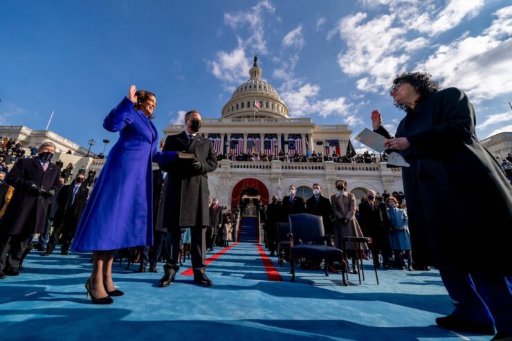 Kamala Harris, in a long blue coat, smiles as she holds up her hand to be sworn in as Vice President. Behind her is the US Capitol, decorated with American flags. Justice Sonia Sotomayor stands several feet away, dressed in her judges robe and swearing Harris in.
